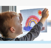Image. A little boy hangs a picture of a rainbow that he created on a window.